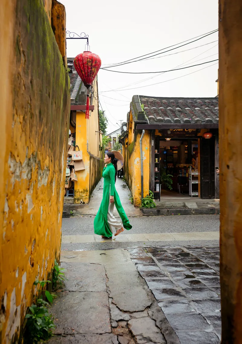 A woman in a green ao dai walking through a narrow street in historic Hoi An