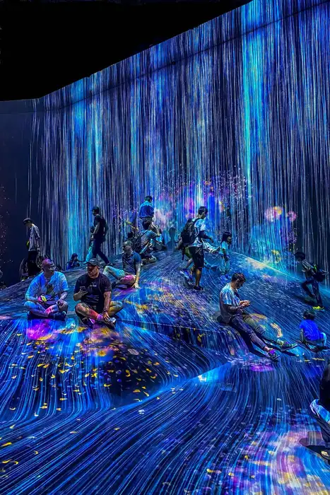 People sitting on a sloped floor inside teamLab Borderless as streams of projected light run down the walls