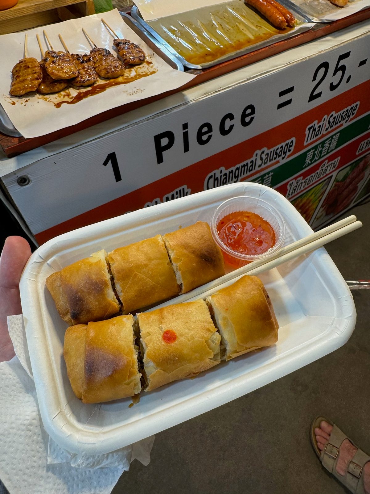 Hand holding a tray of spring rolls and grilled skewers at a street food stall